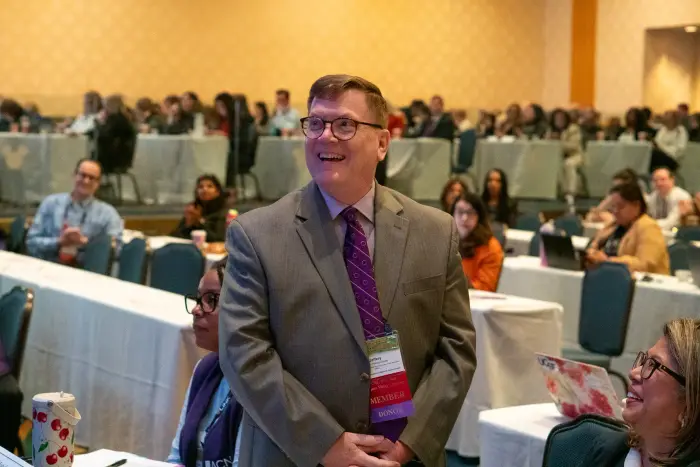A smiling presenter in a suit at the front of a session room, attendees seated behind